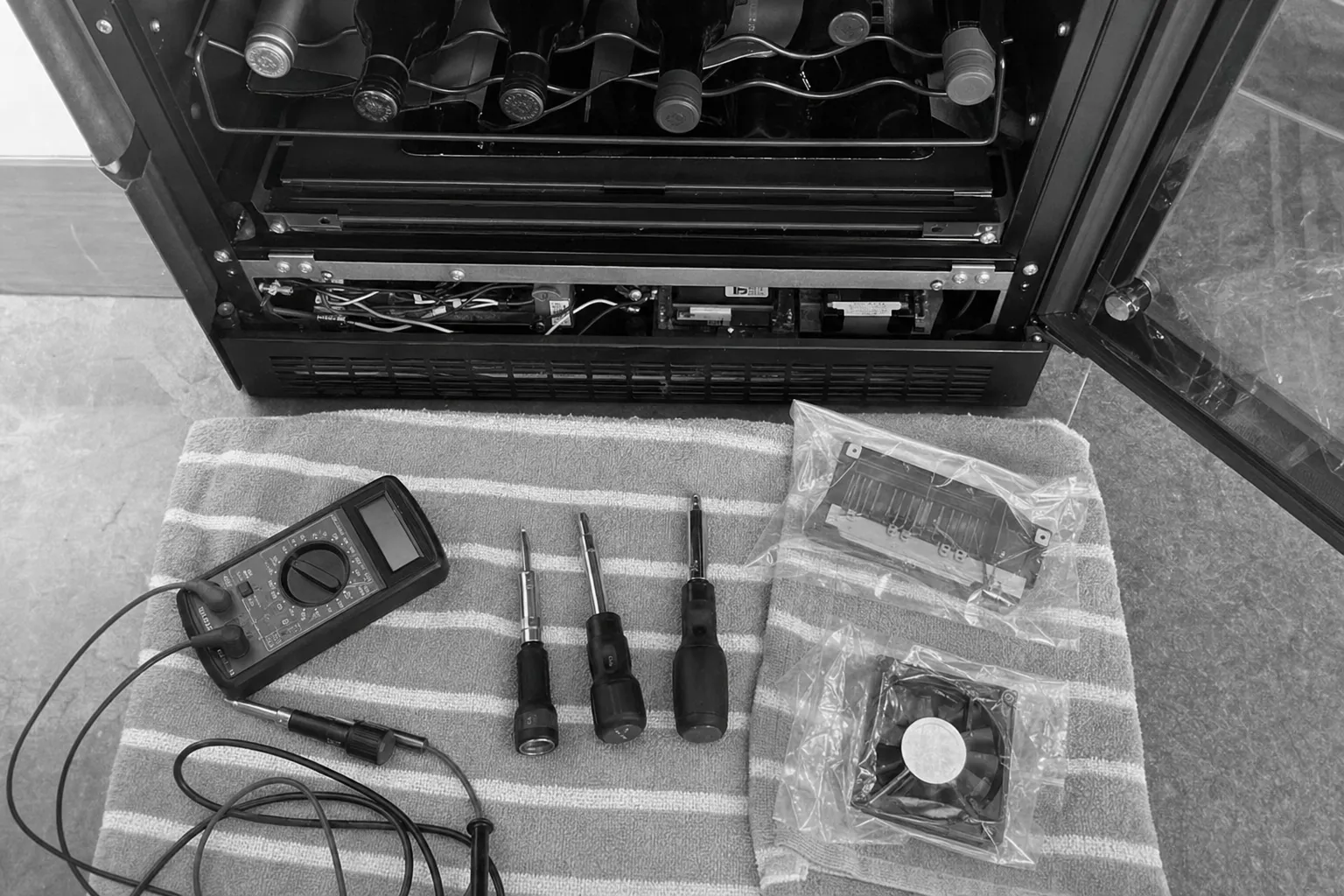 Black-and-white view of a multimeter, three nut drivers, a bagged control board and a bagged fan in front of an opened wine cabinet